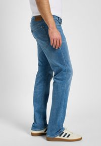 Ljusblå denimjeans med rak benform, med en brun läderpatch vid midjan och kontrasterande sömdekoration.