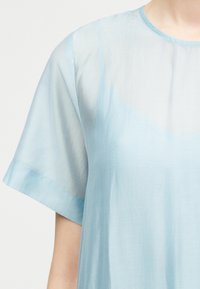 Διαφανές γαλάζιο μπλουζάκι με κοντά μανίκια, φορεμένο πάνω από ένα αντίστοιχο γαλάζιο τοπ, εμφανίζεται από το στήθος μέχρι το μέσο του βραχίονα σε ουδέτερο φόντο.