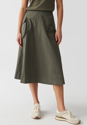 Grøn, mellemlang nederdel lavet af glat stof, med store sidelommer og en tætsiddende talje, kombineret med lyse sneakers.