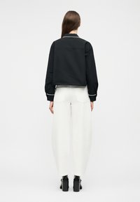 Mujer de cabello largo y castaño que lleva una chaqueta negra con puños y cuello con ribete blanco, pantalones anchos blancos y zapatos de tacón negros, mostrada de espaldas.