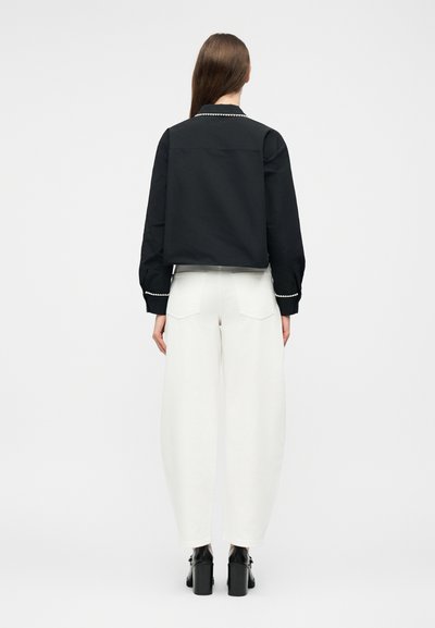 Γυναίκα με μακριά καστανά μαλλιά που φοράει μαύρο μπουφάν με λευκές διακοσμητικές ρίγες στα μανίκια και το γιακά, φαρδιά λευκά παντελόνια και μαύρα παπούτσια με τακούνι, εμφανιζόμενη από την πίσω όψη.