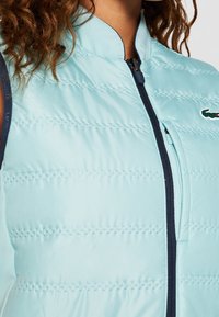 Vaaleansininen toppatakki, jossa on vaakasaumaat, korkea kaulus ja tummansininen vetskari, sisältää Lacoste-logon vasemmalla puolella.