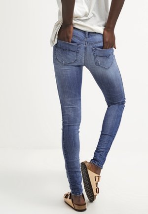 Osoba ubrana w dopasowane niebieskie dżinsy i beżowe sandały typu slide, stojąca ze rękami w tylnych kieszeniach na jednolitym białym tle.