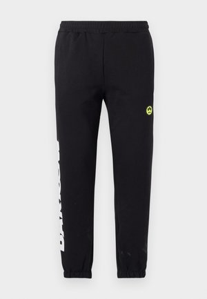 Sorte joggebukser med elastisk midje, smale ben, hvit trykt tekst på siden, og en liten gul smiley-logo på hoften.