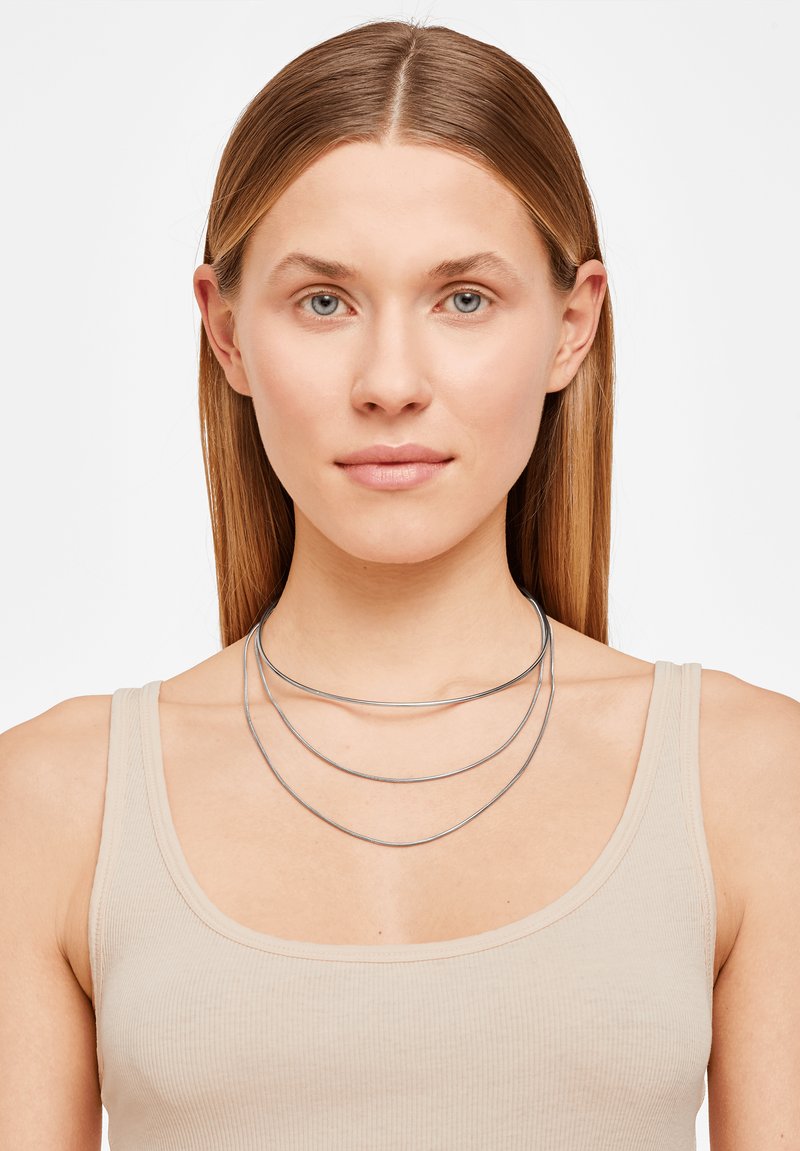 Collier en argent superposé avec trois brins incurvés, présentant une finition métallique lisse, porté sur un débardeur côtelé de couleur claire.