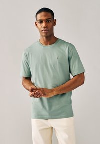 Hellgrünes Baumwoll-T-Shirt mit Rundhalsausschnitt und kurzen Ärmeln, mit einem kleinen bestickten Motiv auf der Brust. Kombiniert mit cremefarbenen Hosen.