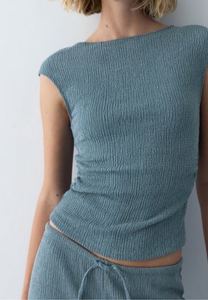 Femme portant un haut sans manches texturé bleu clair et un pantalon à cordon assorti, sur un fond clair uni.