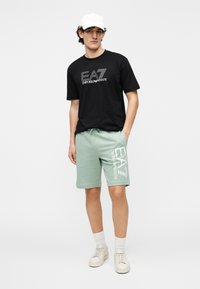 Zwart katoenen T-shirt met wit "EA7 EMPORIO ARMANI" logo, mintgroene shorts met groot wit logo, witte pet, beige sneakers, witte sokken.
