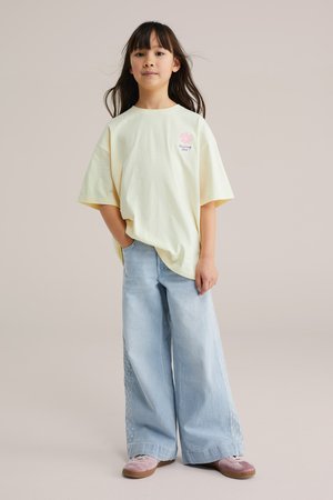 Noor tüdruk, kellel on pikad tumepruunid juuksed, kandes üle suurendatud helekollast t-särki, laiade heleroheliste teksapükste ja roosade kingadega, seisab ühe käega taskus.