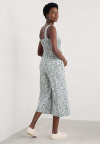 Mono con estampado floral en verde y blanco, cuenta con piernas anchas, cuerpo ajustado y tirantes ajustables; combinado con zapatillas de color claro.