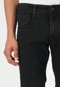Black denim jeans feature a slim fit, subtle whiskering, and a standard five-pocket design with a distinctive back pocket stitch pattern.