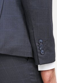 Chaqueta de traje gris oscuro con un sutil patrón de cuadros, tela texturizada y cuatro botones en el puño de un color contrastante.