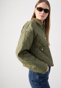 Jachetă cu quilt, de culoare verde măsliniu, cu guler, nasturi cu capse și buzunar frontal; are un design scurt și un material texturat. Potrivită pentru vreme răcoroasă.