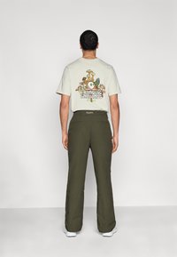 T-shirt clair avec un motif de champignon et le texte "Foraging Fairways" au dos. Pantalon vert olive à coupe décontractée avec des poches.