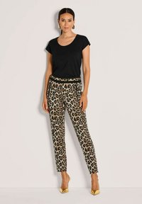 Frau mit schwarzem kurzärmligen Oberteil, Leopardenmuster-Hose mit schwarzem Bund, goldenen spitzen Absätzen und goldenen geometrischen Ohrringen, stehend.