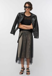 Giacca da motociclista in pelle nera indossata sopra una maglia nera trasparente, abbinata a una lunga gonna traslucida e tacchi neri con cinturini e dettagli argentati.