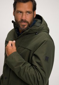 Olijfgroene geïsoleerde jas met een capuchon, voorzien van een ritsluiting, voorksam en een gestructureerd label op de linkermouw.
