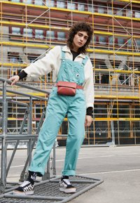Pantalones de tirantes verde menta de algodón, con un bolsillo frontal, combinados con un suéter de felpa blanco, calcetas negras con estampado y zapatos de cuadros.