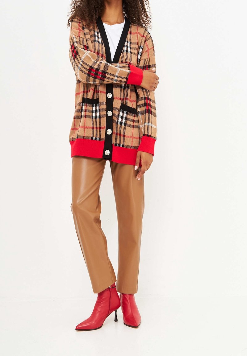 Cardigan a cuadros beige con acentos negros, puños rojos y grandes botones, combinado con pantalones de piel sintética color beige y botas de tacón puntiagudas rojas.