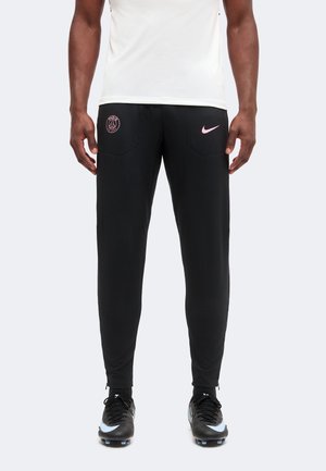 Homme portant un jogging noir Paris Saint-Germain Nike avec des logos roses et des crampons noirs, debout devant un fond uni.