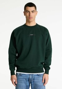 Joven usando un sudadera verde oscura con texto tenue y jeans azul claro, de pie contra un fondo blanco sencillo.