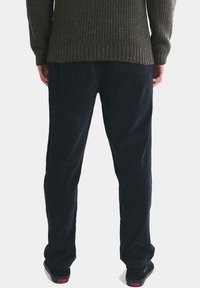 Pantalones de pana negra con un corte relajado y textura ribeteada vertical, combinados con un suéter de punto verde oscuro. Zapatos oscuros visibles.