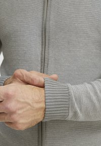 Szürke textúrájú zipzáras kardigán világos és sötét csíkokkal, bordás mandzsettákkal és normál fazonnal, kiemelve az anyag kötési mintázatát.