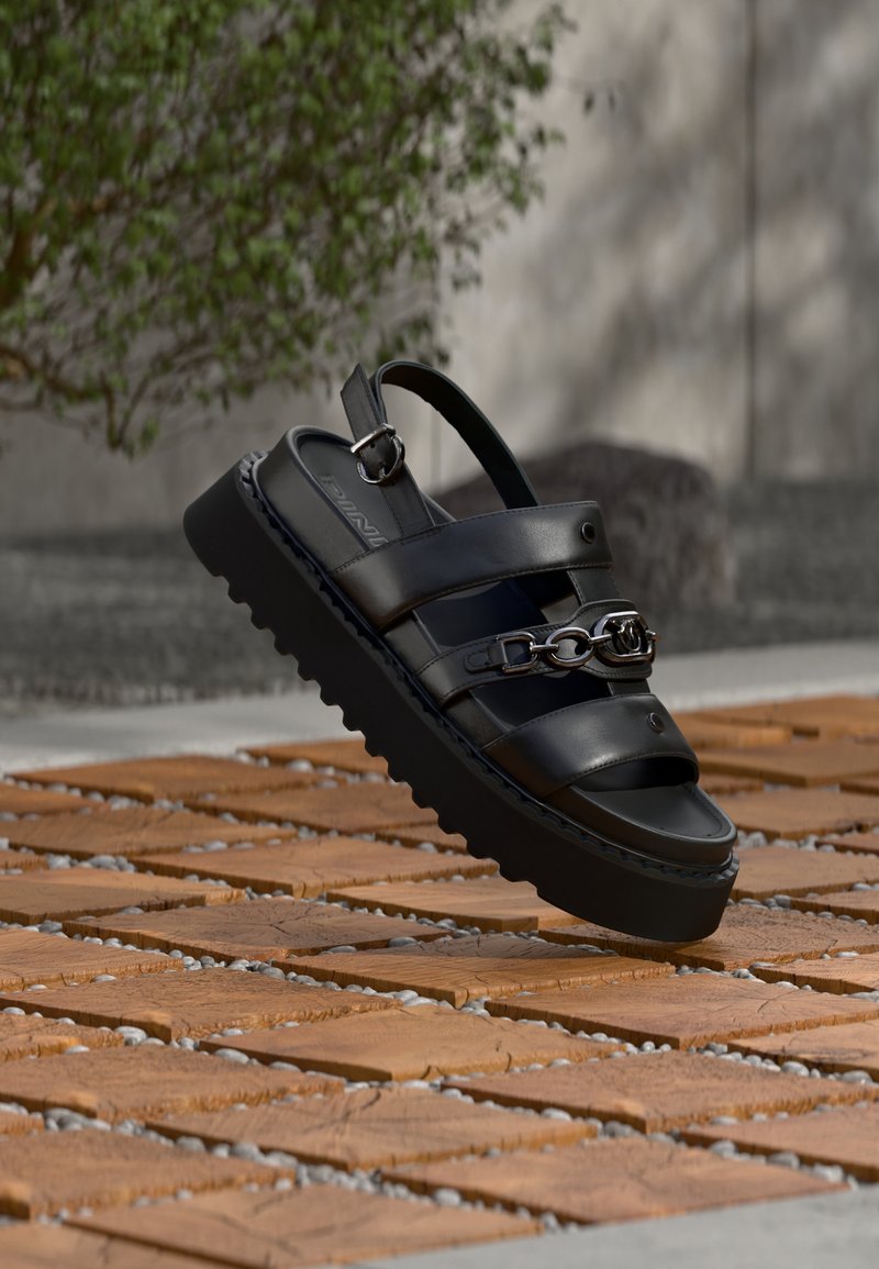 Μαύρο πλατφόρμα σανδάλι με τρεις λουριά, μεταλλική αλυσίδα και αγκράφα, σε καφέ πλακόστρωτη επιφάνεια σε εξωτερικό χώρο με θολό πράσινο φόντο φυτού.