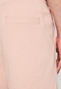 Gros plan sur un pantalon en tissu tricoté beige montrant une poche passepoilée arrière et la main droite d'une personne reposant à côté de la hanche.