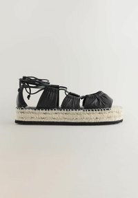 Črne usnjene espadrile s pleteno juto platformo. Značilnosti vključujejo mehko usnjeno nabiranje in nastavljive veze okoli gležnja za varno prileganje.