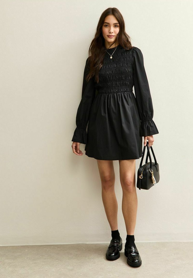 Czarna sukienka z marszczonym gorsetem, długimi bufiastymi rękawami i rozszerzanymi mankietami. W połączeniu z elegancką czarną torebką oraz błyszczącymi loafersami.