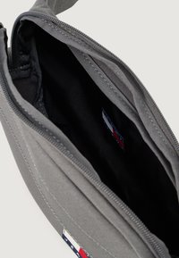 Borsa di tessuto grigia con chiusura a zip, interni rivestiti in nero, presenta un piccolo etichetta con logo in accenti rossi, bianchi e blu.