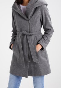 Šedý kabát z vlněné směsi s kapucí, zapínáním na zip, páskem na zavázání a bočními kapsami. Hladká textura s přizpůsobeným střihem a jemnými šitími detaily.