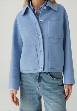 Personne portant une veste en laine bleu clair boutonnée avec trois poches à l'avant, par-dessus une chemise rayée bleue et un jean bleu délavé.