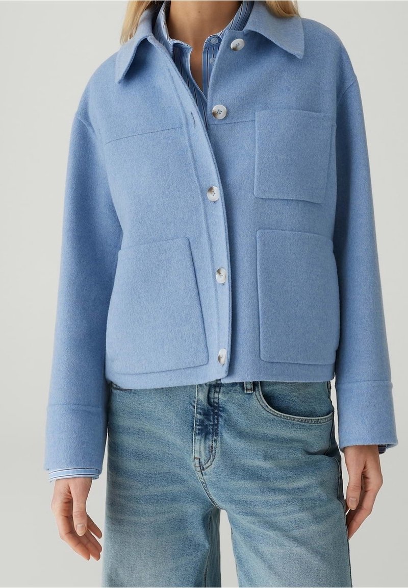 Personne portant une veste en laine bleu clair boutonnée avec trois poches à l'avant, par-dessus une chemise rayée bleue et un jean bleu délavé.