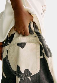Jeans camo in beige e nero con un tessuto denim testurizzato. Il primo piano mostra una mano nella tasca e dettagli di cucitura visibili.