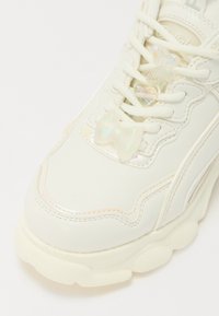 Adidași albi din material sintetic lucios, cu vârful rotunjit, talpă groasă și farmecuri decorative în formă de fluturi și fundițe.