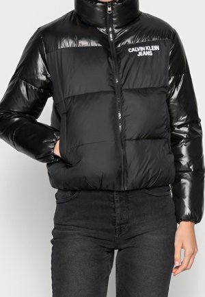 Czarna kurtka puchowa z błyszczącymi rękawami, zamkiem z przodu, bocznymi kieszeniami oraz białym logo "Calvin Klein Jeans" na piersi, noszona z czarnymi jeansami.