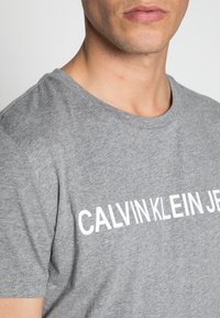 Camiseta de algodón de manga corta en color gris, que presenta un gran logo blanco "CALVIN KLEIN JEANS" en el pecho. Textura suave.