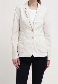 Vaaleanbeige bleiseri, jossa on jämäkkä muotoilu, kaksi nappia, kauluksen läppä, etutaskut ja sileä pinta, pukeutuneena valkoisen paidan päälle.
