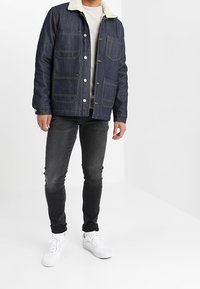 Chaqueta de mezclilla con un cuello forrado de piel, cuatro bolsillos frontales, tejido azul, costuras marrones, combinada con pantalones vaqueros negros y zapatillas blancas.