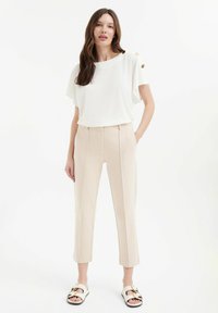 Witte top met korte mouwen en gouden knoopaccenten, beige op maat gemaakte pantalon met rechte pijpen, en witte sandalen met zwarte accenten en gouden hardware.