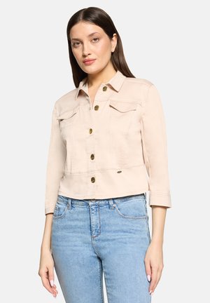Woman wearing light beige cropped jacket with gold buttons and light blue high-waisted jeans, standing against a white background.