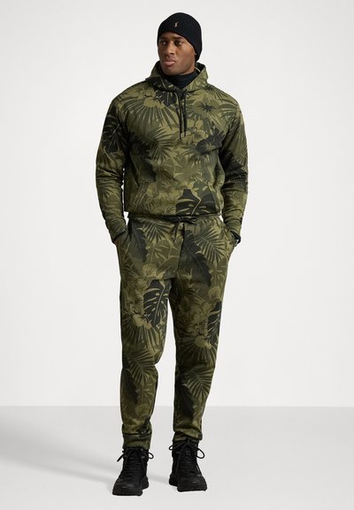 Olivově zelená mikina s kapucí a odpovídající kalhoty s botanickým potiskem. Má kapsy a stahovací pás. Černá obuv.
