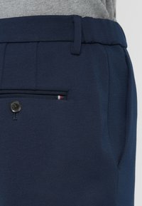 Záber na tmavomodré nohavice so zašnurovanou zadnou kapsou, pútkom na opasok a časťou šedého pleteného svetra zasunutého vo vnútri.