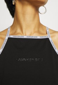 Närbild av en person som bär en svart halterneck-top från Calvin Klein Jeans med vita logoband och silverfärgade hoopsörhängen.