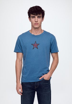 Un jeune homme porte un tee-shirt bleu orné d’un motif représentant une étoile rouge et un jean foncé, sur un fond blanc uni.