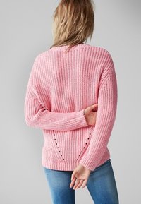 Różowy sweter z dzianiny o ribbowym wykończeniu, z opadającymi ramionami i szczegółowymi wycięciami wzdłuż bocznych szwów. W zestawie z niebieskimi dżinsami.