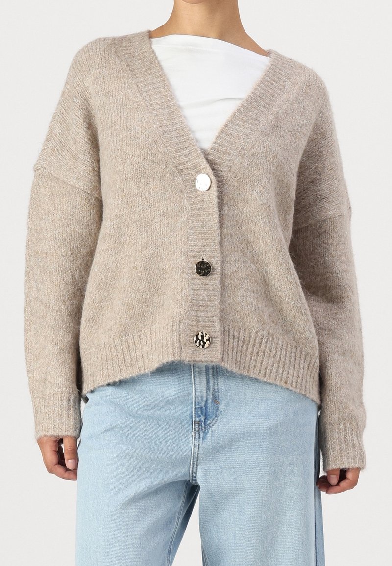 Cardigan beige en maille épaisse avec un col en V profond et trois boutons dépareillés, porté sur un haut blanc et associé à un jean bleu clair.