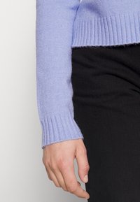 Suéter de punto de color púrpura claro y acanalado, con mangas largas, combinado con tejido negro. Primer plano de una mano descansando en la cintura.